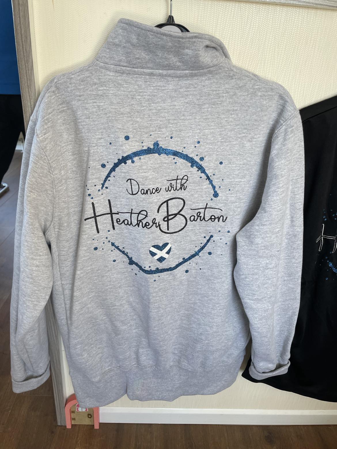 a photo of a grey jacket showing the Dance with Heather Barton in sparkly blue and black glitter on the back